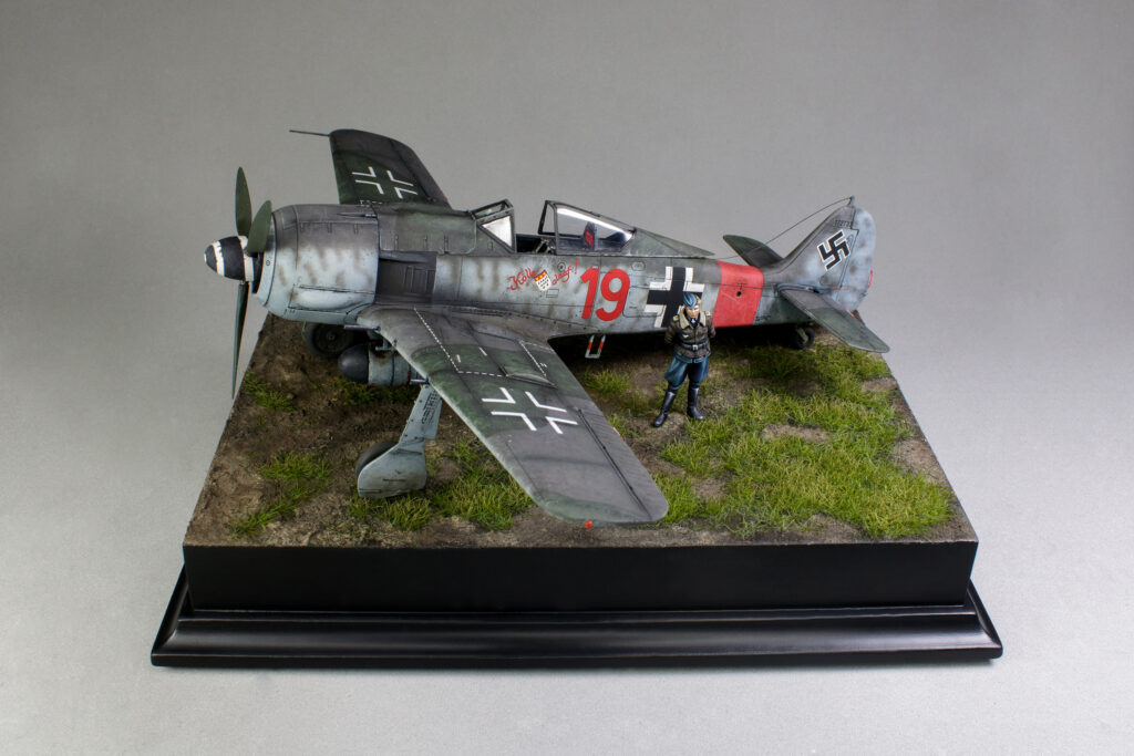 Side view of the model of a Focke Wulf FW 190 A8 in mid-war camouflage. The aircraft is on a diorama decorated with green grass. Pilot Ernst Schroder poses proudly in front of it, dressed in an aviator jacket.